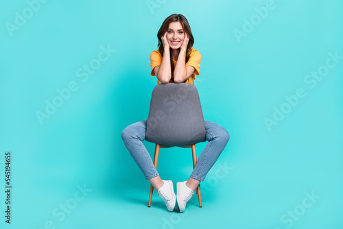 Full length photo of pretty sweet girl dressed yellow t-shirt sitting chair smiling arms cheeks isolated teal color background