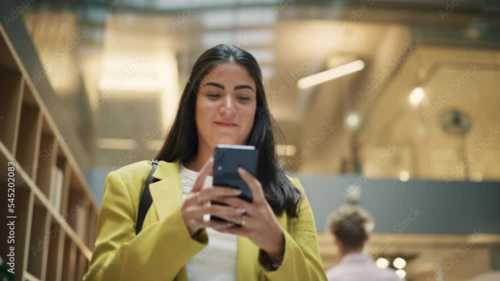 Portrait of a Happy Hispanic Woman Texting on her Smartphone and Walking in Corporate Office. Female Marketing Manager Checking Social Media Trends and Staying Updated. Low Angle Slow Motion Portrait of a Happy Hispanic Woman Texting on her Smartphone and Walking in Corporate Office. Female Marketing Manager Checking Social Media Trends and Staying Updated. Low Angle Slow Motion