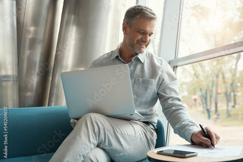 Gray-haired man is comfortably seated with laptop and documents on sofa