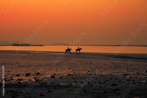Beautiful calm orange sunset over the coastline with horse riders © Ebster/Wirestock Creators