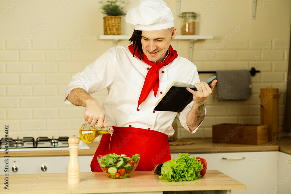 Young man dressed as chef pouring olive oil on vegetable salad, reading recipe book in kitchen. Concept of food, cooking, healthy lifestyle. Young man dressed as chef pouring olive oil on vegetable salad, reading recipe book in kitchen. Concept of food, cooking, healthy lifestyle.