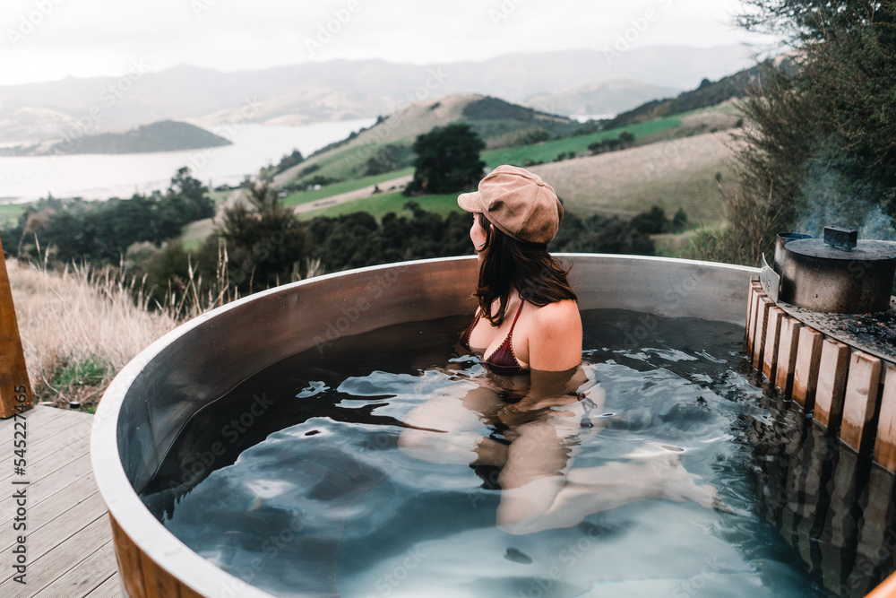 caucasian girl wearing swimsuit and brown hat inside round hot water jacuzzi looking at beautiful landscape of french farm bay calm sitting and relaxing near mountains and peaceful nature, te wepu - obrazy, fototapety, plakaty 