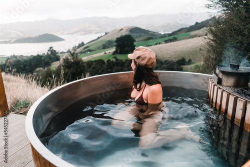 caucasian girl wearing swimsuit and brown hat inside round hot water jacuzzi looking at beautiful landscape of french farm bay calm sitting and relaxing near mountains and peaceful nature, te wepu