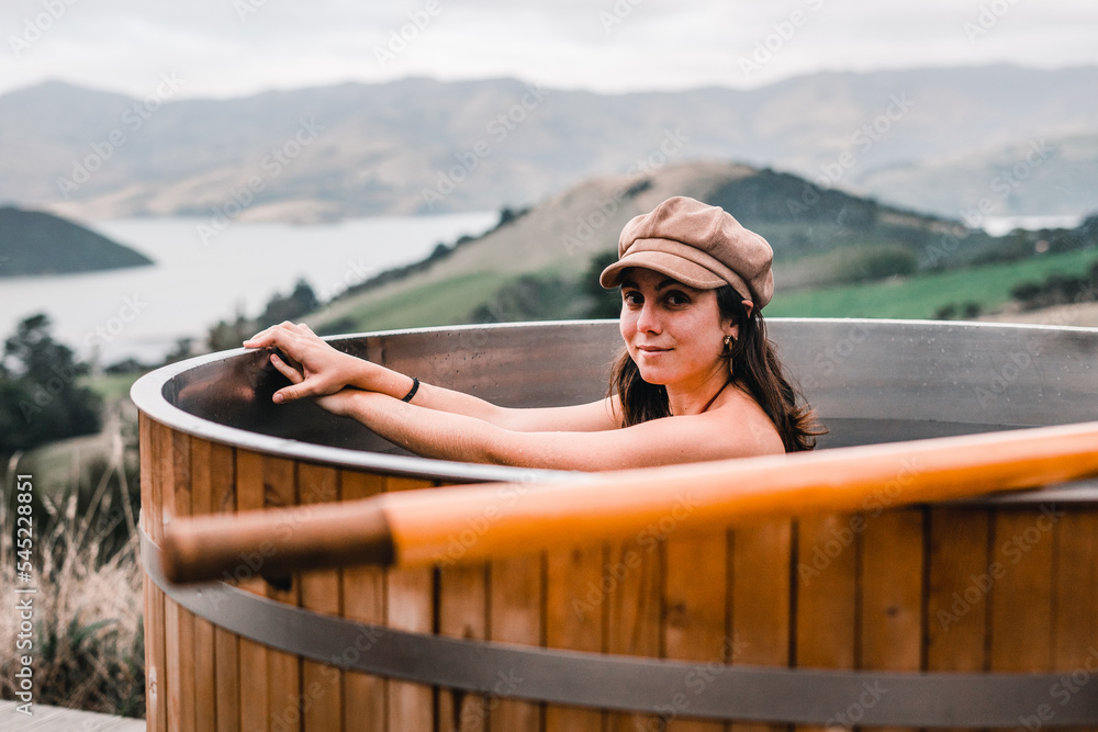 blue-eyed caucasian young girl with brown hat and bracelet on her wrist and crossed hands inside the hot water of the round jacuzzi in the middle of the vegetation and mountains, te wepu pods akaroa