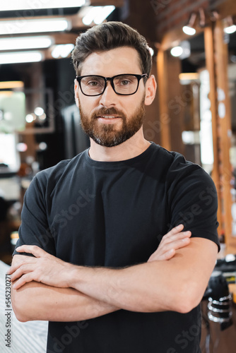Bearded barber in eyeglasses crossing arms and looking at camera in barbershop.