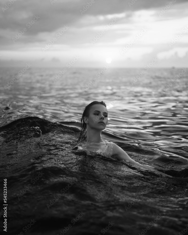 Fototapeta premium Black and white snapshot of a beautiful woman in the ocean. Dark portrait of a sensual gentle girl in the water in summer. Brunette woman is swimming and relaxing in the water before sunset. 