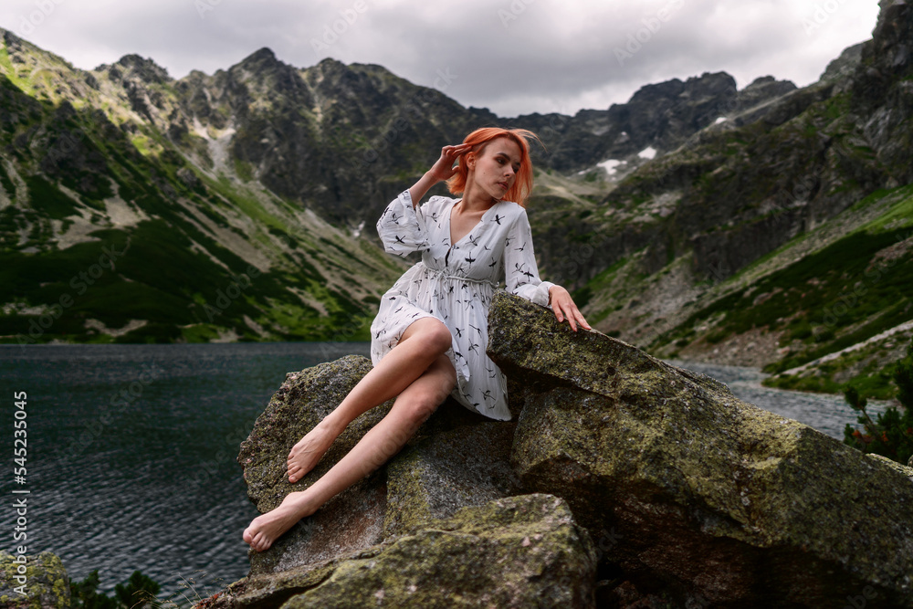 Naklejka premium Elegant sexy lady with red hair in a white dress sits on the stone in the mountains and relaxes under sun rays. Fashion natural portrait as wallpaper, magazine design or advertising cover.