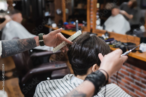 tattooed barber with comb and blurred scissors near brunette teenager in beauty salon.