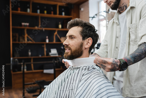 tattooed barber wrapping neck of bearded man with hairdressing collar.