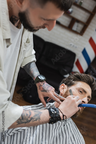 blurred hairdresser in leather bracelet shaving cheek of man with straight leather.