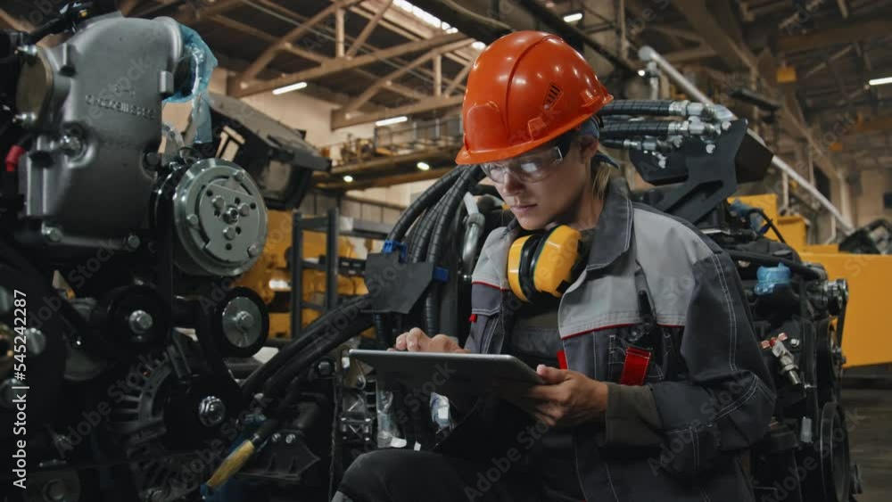 Young female engineer in safety glasses and hard hat taking notes on digital tablet while examining modern machine equipment at industrial plant Young female engineer in safety glasses and hard hat taking notes on digital tablet while examining modern machine equipment at industrial plant