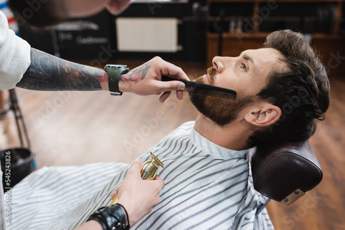 tattooed barber holding hair clipper and combing beard of man in barbershop.