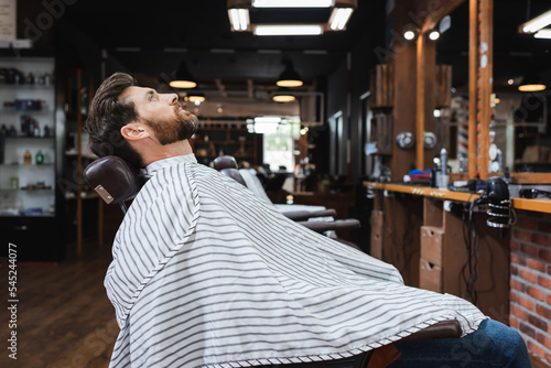 side view of brunette bearded man sitting in hairdressing cape in beauty salon.