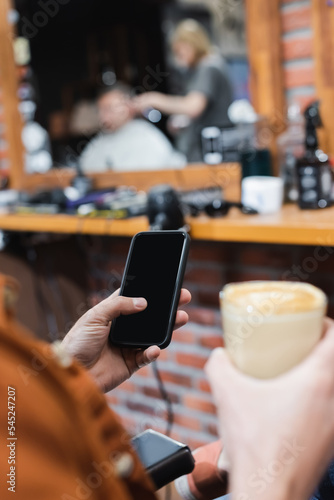 cropped view of blurred man holding smartphone with blank screen and glass of cappuccino in barbershop.