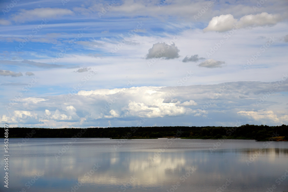 Obraz premium July. Blue sky and white clouds. Forest lake and reflection in the water. The banks of the reservoir, 