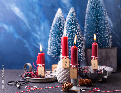 Christmas composition. Red burning candles with boardes with numbers, cones,  wild blue berries and blue decorative trees against blue  textured background. Scandinavian minimalistic style. Still life photo