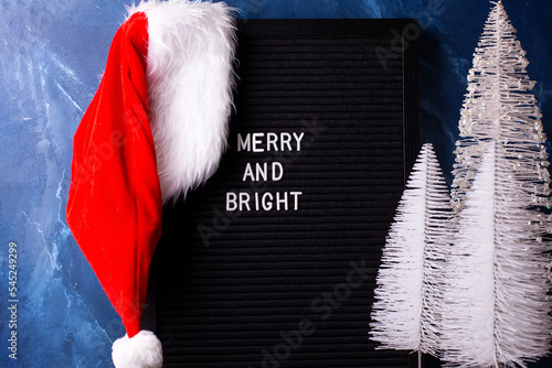 Winter holidays decorations. Black board, red hat of Santa Claus and b white Christmas trees against deep blue marble wall.   Still life. Christmas concept. Winter holiday postcard..