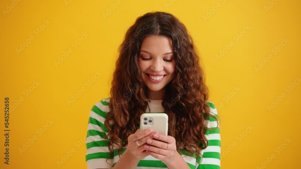 Pensive curly haired brunette woman typing on mobile in the yellow studio Pensive curly haired brunette woman typing on mobile in the yellow studio