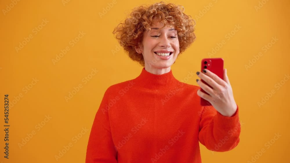 Pretty curly haired adult ginger woman talking by video call on phone and greeting in the yellow studio Pretty curly haired adult ginger woman talking by video call on phone and greeting in the yellow studio