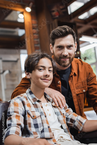 Teen boy looking at camera near blurred hairstylist in beauty salon.