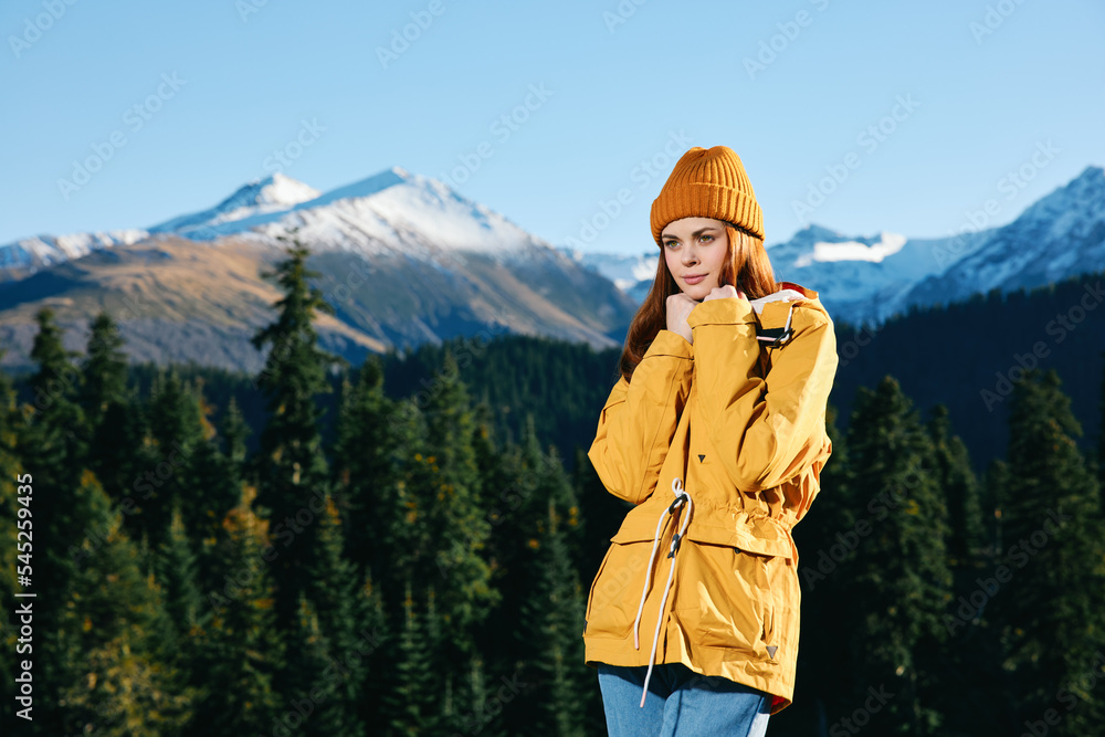 Woman with red hair hiker standing on the mountain hands up happiness overlooking snowy mountains and trees in yellow raincoat and cap travel autumn and hiking in the mountains in the sunset freedom