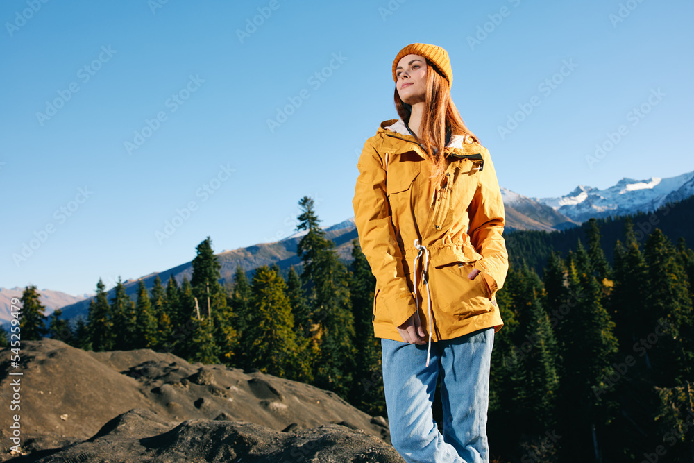 Woman with red hair hiker in yellow raincoat and cap standing on a mountain overlooking snowy mountains traveling in winter and hiking in the mountains in the wilderness at sunset Woman with red hair hiker in yellow raincoat and cap standing on a mountain overlooking snowy mountains traveling in winter and hiking in the mountains in the wilderness at sunset