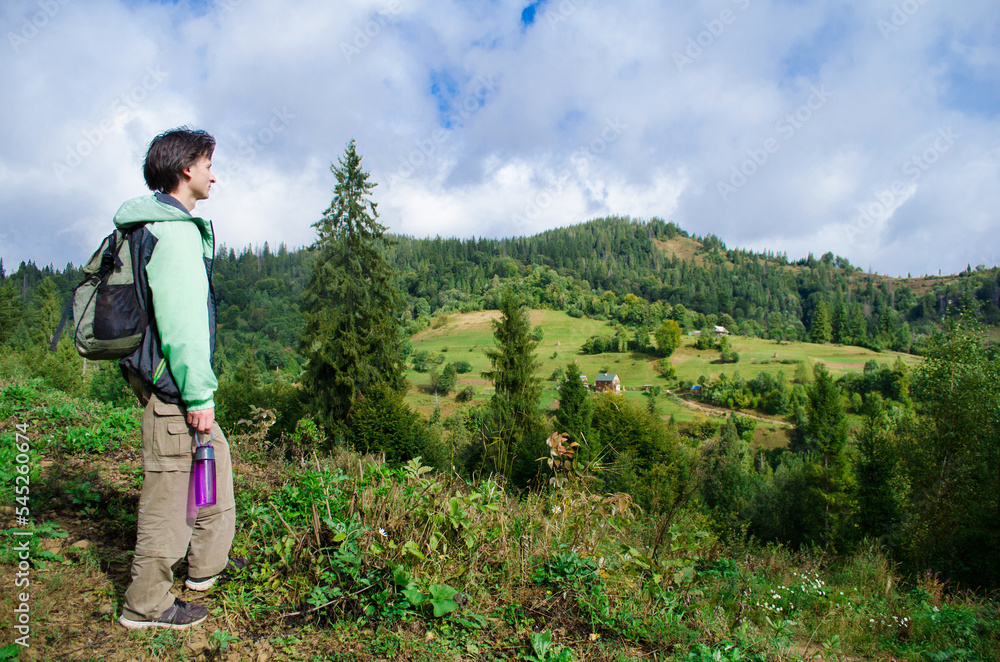 Naklejka premium Young man hiker in a green jacket and with a backpack looks at the autumn mountains with a forest, photo of a tourist