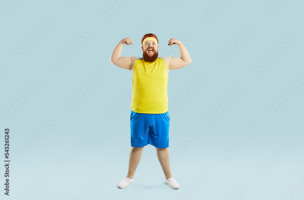 Funny happy positive cheerful excited motivated fat man in sweatband, yellow top and blue shorts smiling, flexing arms and showing his weak muscles. Sports exercise, fitness workout at the gym concept Funny happy positive cheerful excited motivated fat man in sweatband, yellow top and blue shorts smiling, flexing arms and showing his weak muscles. Sports exercise, fitness workout at the gym concept