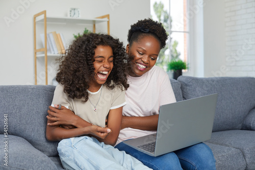 Happy family watching something funny on laptop. Cheerful young African American mother and daughter sitting on sofa at home, looking at notebook computer and laughing together