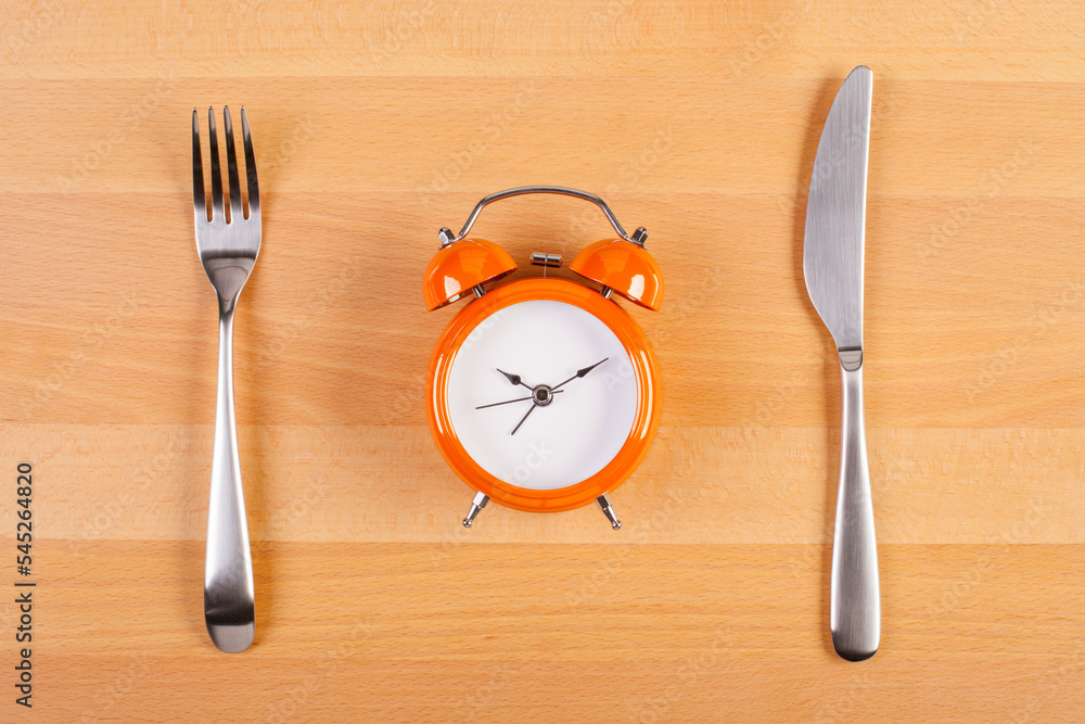 Naklejka premium alarm clock on a plate that stands between fork and knife on a wooden table. flat lay time to eat concept image