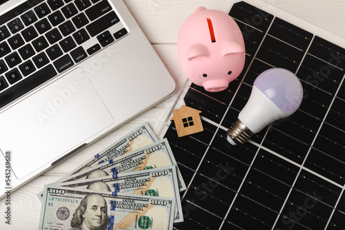 Flat lay composition with solar panel, led lamp, house model and money on  table. Money saving and clean energy concept. Ecology and sustainable development concept. photo