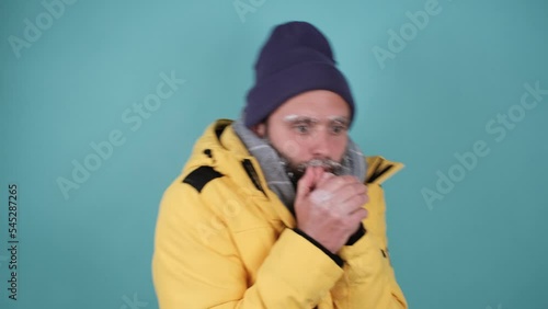 The frozen man is shivering from the cold, a frozen face covered with frost, he wears a jacket with a hood and is wrapped in a warm scarf isolated on a blue background .Energy crisis concept in Europe