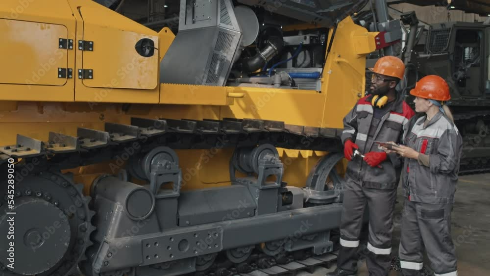 Two diverse tractor mechanics in coveralls inspecting tractor machine standing indoors of manufacturing plant Two diverse tractor mechanics in coveralls inspecting tractor machine standing indoors of manufacturing plant