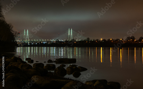 Night warm cloudy landscape of the river bank, rocky coast, night city lights, the Bolshoy Obukhovsky Bridge (cable-stayed bridge) across the Neva river.