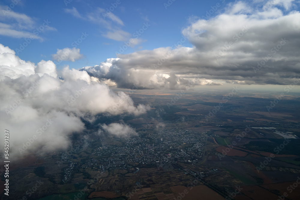 Fototapeta premium Aerial view from airplane window at high altitude of distant city covered with puffy cumulus clouds forming before rainstorm