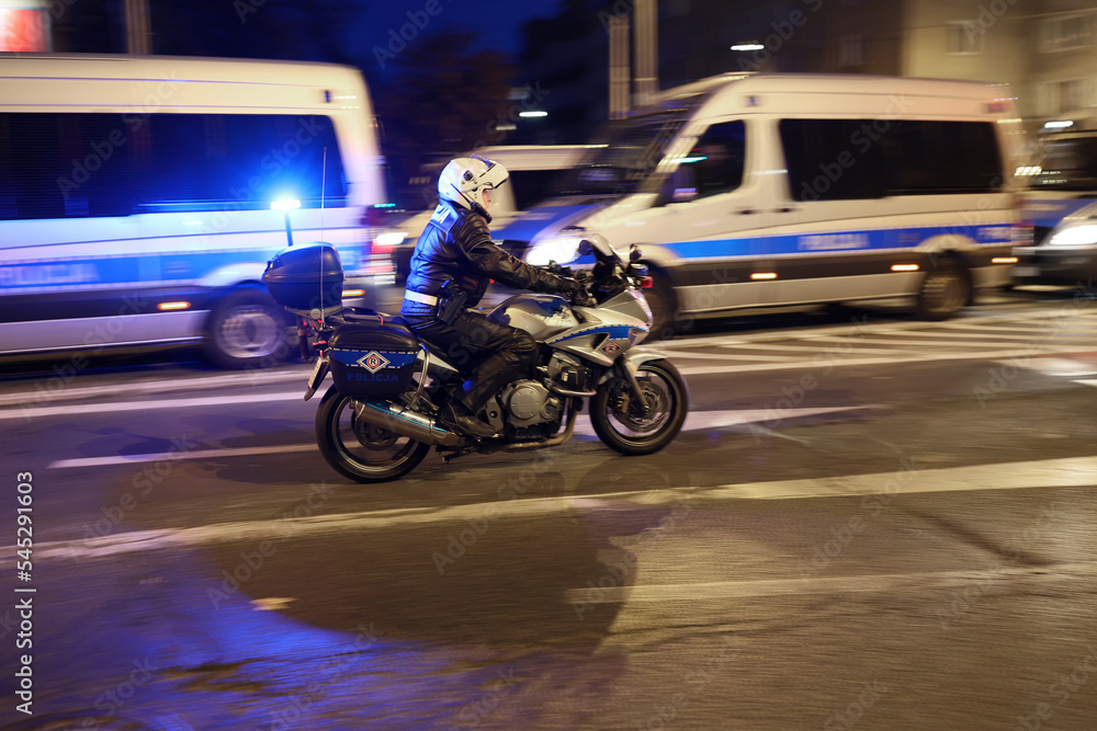 Policjant wydziału ruchu drogowego z motocyklem podczas kontroli miasta wieczorem.  Światła policyjne. - obrazy, fototapety, plakaty 