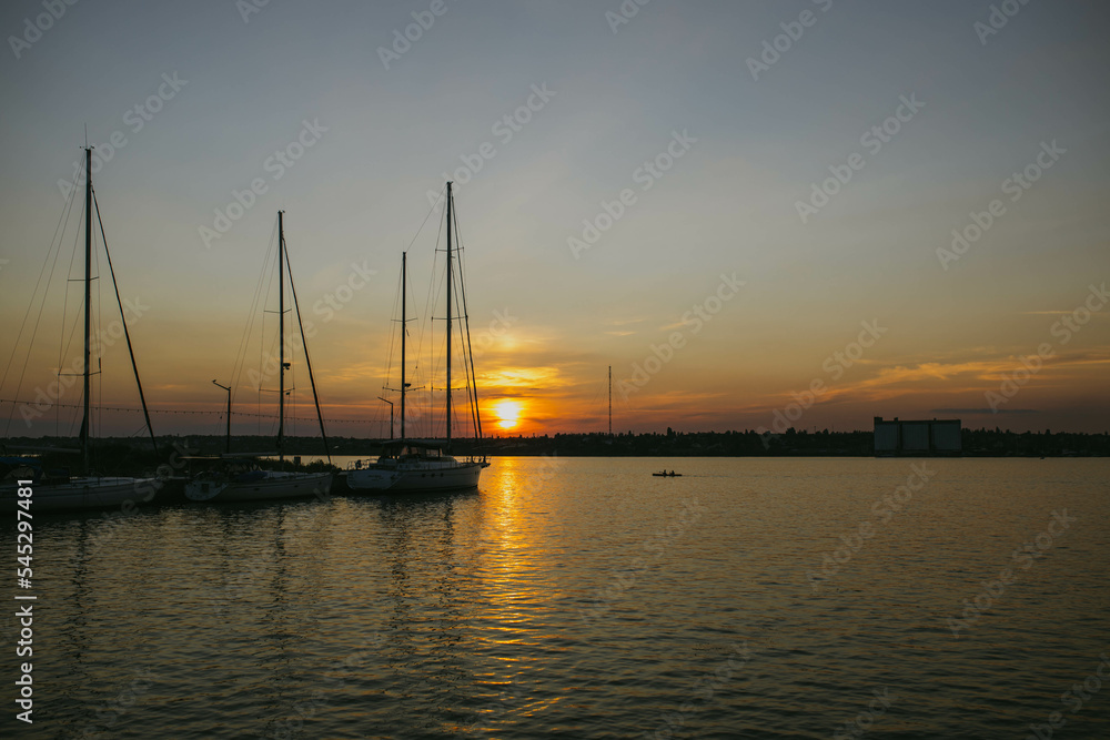South Ukraine, Mykolaiv - August 21, 2021: Sunset in the river coast side, sun reflects into the water surface. Yachts and boats near the beach, people resting, Calm atmosphere in peaceful  city