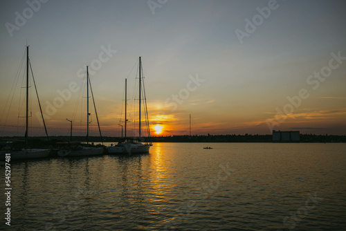 South Ukraine, Mykolaiv - August 21, 2021: Sunset in the river coast side, sun reflects into the water surface. Yachts and boats near the beach, people resting, Calm atmosphere in peaceful city