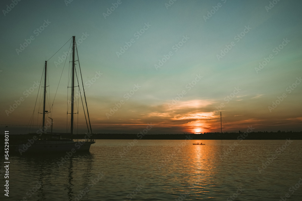 South Ukraine, Mykolaiv - August 21, 2021: Sunset in the river coast side, sun reflects into the water surface. Yachts and boats near the beach, people resting, Calm atmosphere in peaceful city South Ukraine, Mykolaiv - August 21, 2021: Sunset in the river coast side, sun reflects into the water surface. Yachts and boats near the beach, people resting, Calm atmosphere in peaceful city