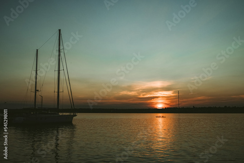 South Ukraine, Mykolaiv - August 21, 2021: Sunset in the river coast side, sun reflects into the water surface. Yachts and boats near the beach, people resting, Calm atmosphere in peaceful  city © Евгения Жигалкина