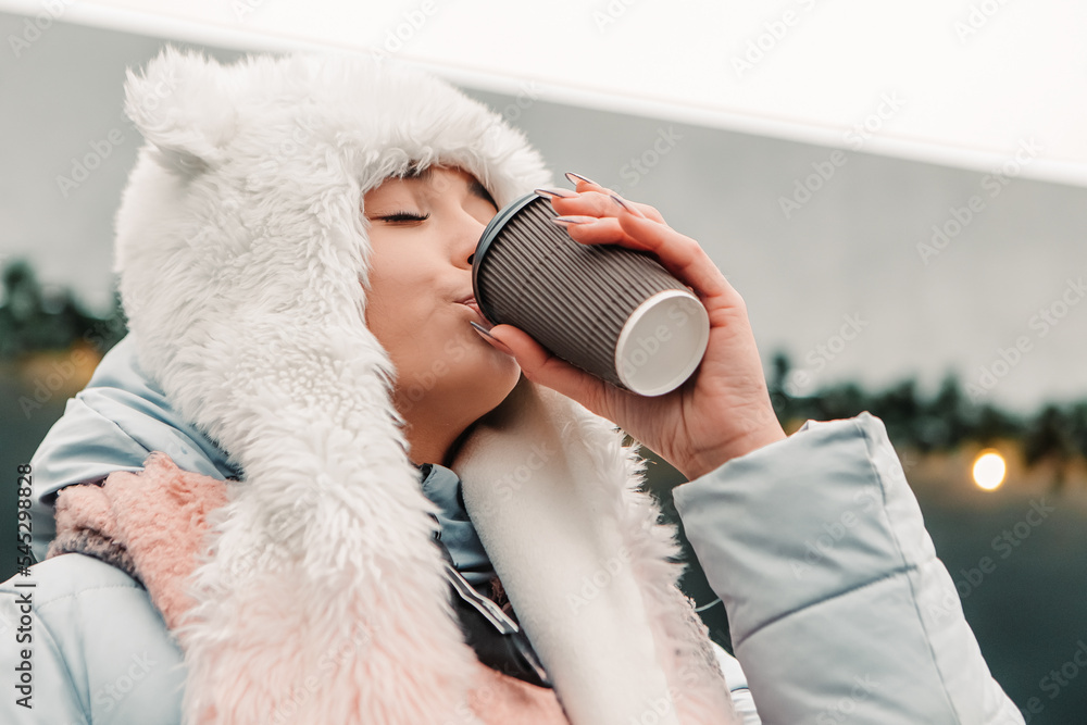 Young pretty smiling woman celebrating dressed warm jacket and woolen hat, drink hot tea coffee beverage mulled wine. Winter holidays, travel spirit resort. New Year Christmas decorations, garlands
Young pretty smiling woman celebrating dressed warm jacket and woolen hat, drink hot tea coffee beverage mulled wine. Winter holidays, travel spirit resort. New Year Christmas decorations, garlands