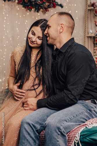 Vertical image of man hug a woman with round belly with baby kid inside sitting near Christmas Tree, kissing, celebrating New Year holiday. Pregnant lady waiting for infant. Expecting a newborn child 