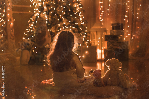 Rear view, little girl sits with plush toys on woolen carpet, looks at Christmas tree with gifts and glowing garland, back to camera against background of Christmas tree. Photo with retro film effect.