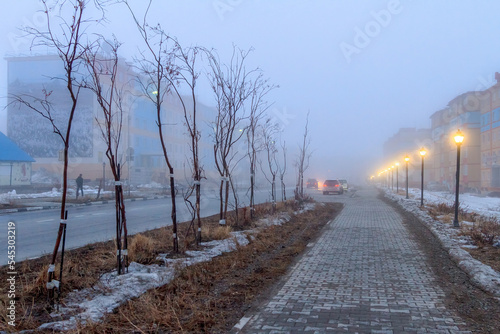 Evening urban landscape. View of the sidewalk and street lights. Mid-May in the Arctic. Snow is melting on the streets of the city. Cold fog. City of Anadyr, Chukotka, Siberia, Russia. © Andrei Stepanov