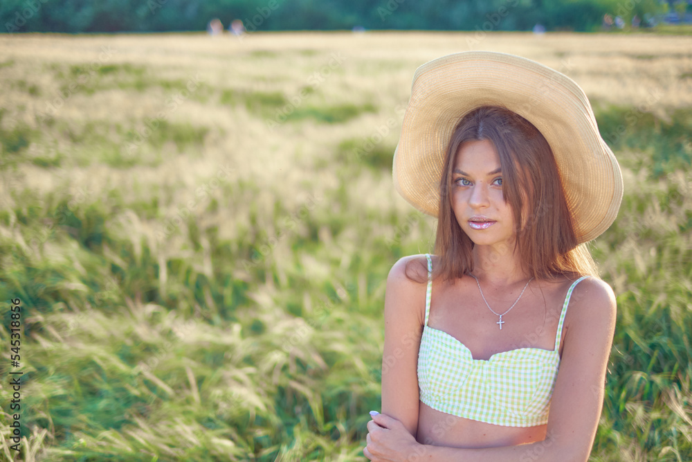 stylish beautiful smiling young girl in a straw hat and a fashionable dress walks around the field and poses