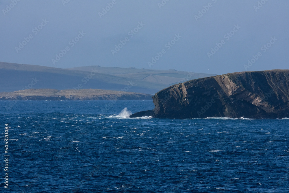 Cliffs and rocky coastline silhouette coast of Shetland Islands in Atlantic Ocean on sunny day with clouds, lighthouse and hills seen from cruiseship cruise ship liner with spray