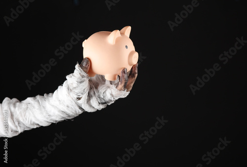 Canvas Print Mummy's hand wrapped in bandage holds piggy bank isolated on black background