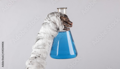 Φωτογραφία Mummy's hand wrapped in a bandage holds a laboratory flask with blue elixir isolated on a black background
