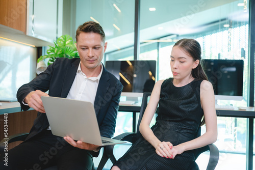 Two young business woman discussing project and report in laptop and digital tablet while standing and analyzing data on statistics and brainstorming information in modern office