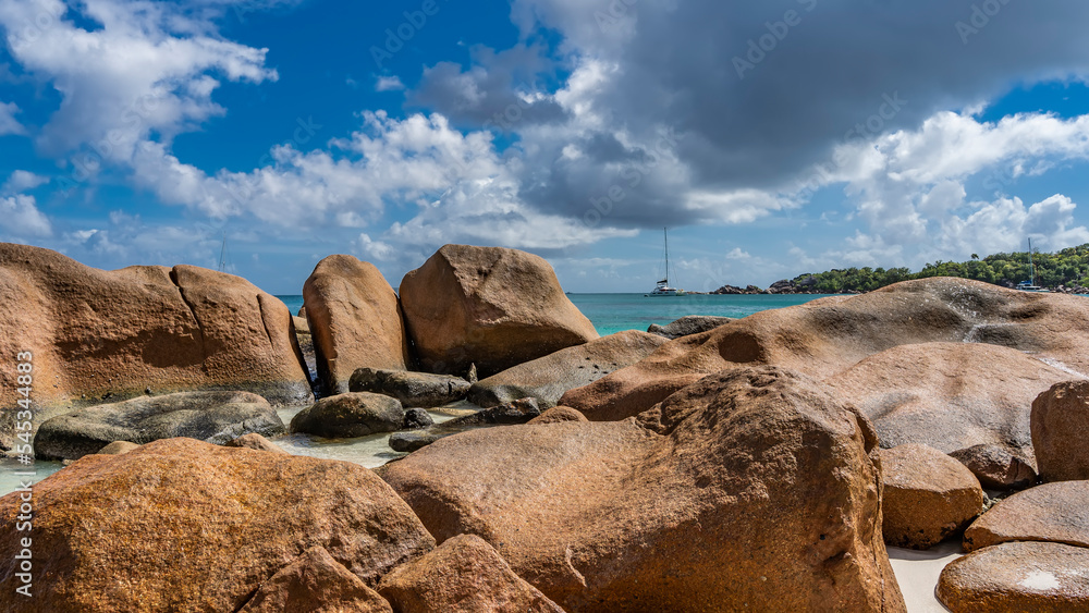 Picturesque granite boulders are piled on the ocean shore. In the distance, on the turquoise water, a yacht is visible. Clouds in the blue sky. Seychelles. Praslin. Anse Lazio beach   